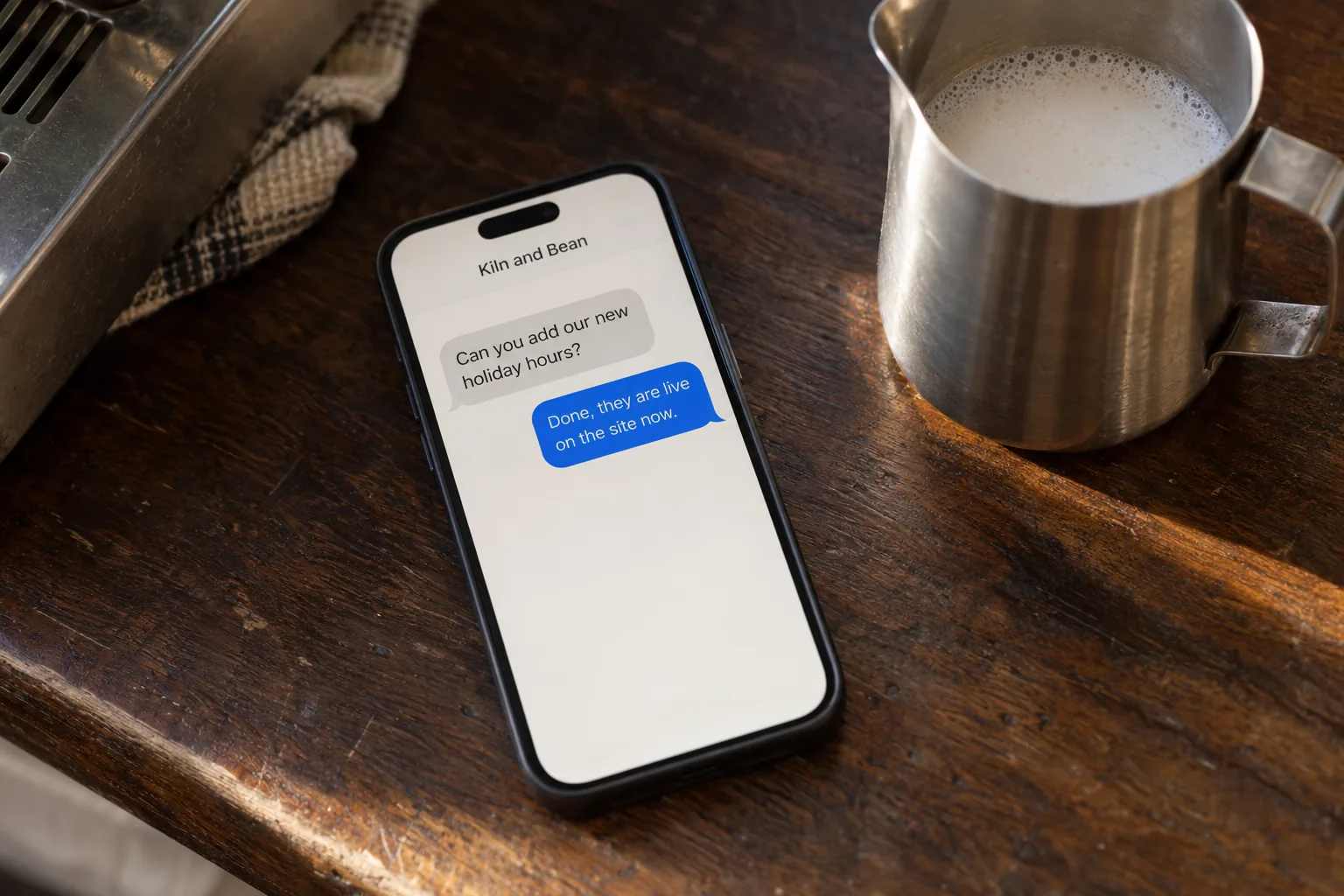 A phone on a cafe counter showing a text thread with Kiln and Bean asking for holiday hours to be added concept